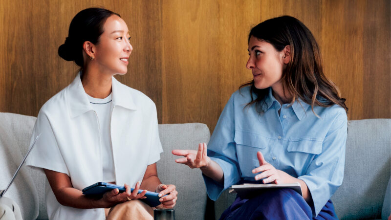 Two women talking and smiling on a sofa, one holding a tablet and the other gesturing with a notebook on her lap.