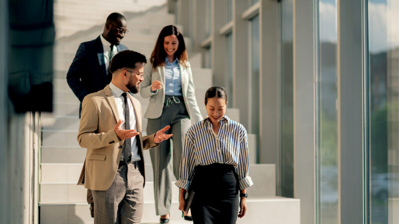 Four colleagues chat and smile while walking down a sunlit outdoor staircase beside a glass building.